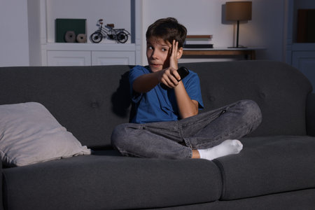 Emotional boy changing TV channels with remote control on sofa at homeの写真素材