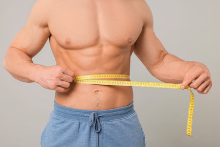Athletic man measuring waist with tape on light gray background, closeup. Weight loss conceptの写真素材