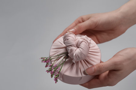 Furoshiki technique. Woman holding gift packed in fabric and beautiful pink flowers on gray...