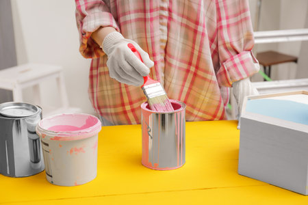 Woman dipping brush into can of pink paint at yellow wooden table indoors, closeupの写真素材