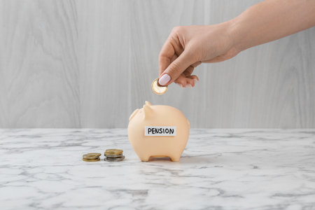 Pension savings. Woman putting coin into piggy bank at white marble table, closeup