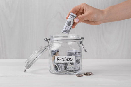 Pension concept. Woman putting dollar banknote into glass jar with money at white wooden table, closeupの写真素材