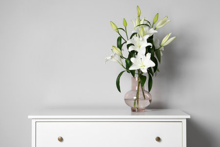 Beautiful bouquet of lily flowers in glass vase on chest of drawers near white wall, space for textの写真素材