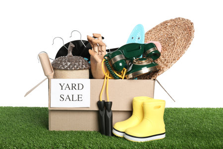 Sign Yard Sale written on box with different stuff on green grass against white background