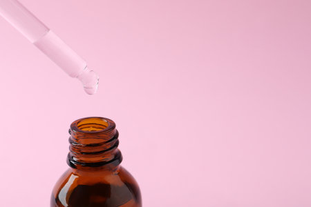 Dropping cosmetic oil from pipette into bottle on pink background, closeup. Space for textの写真素材