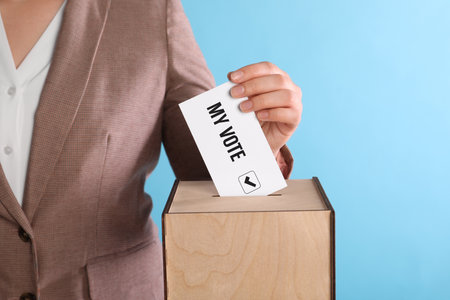 Woman putting paper with text My Vote and tick into ballot box on light blue background