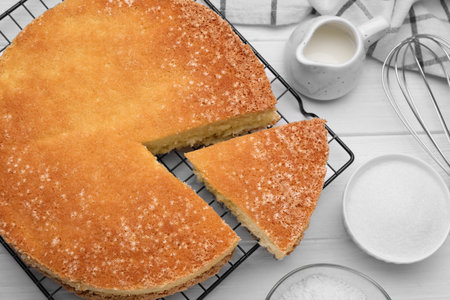 Tasty sponge cake, whiskey and ingredients on white wooden table, flat lay