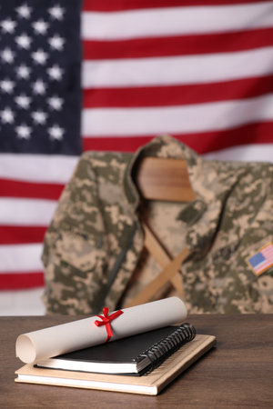 Notebooks and diploma on wooden table, chair with soldier uniform against flag of United States indoors. Military educationの写真素材