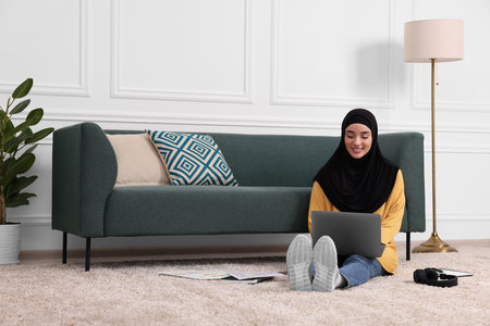 Muslim woman in hijab using laptop on floor near sofa in room. Space for textの写真素材