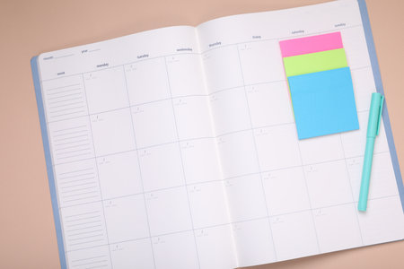 Open monthly planner, blank sticky notes and pen on beige background, top view. Space for text