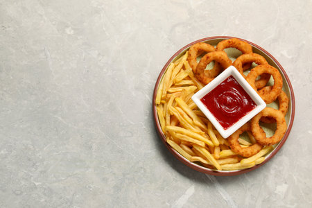 Plate with tasty ketchup, fries and onion rings on light gray table, top view. Space for textの写真素材