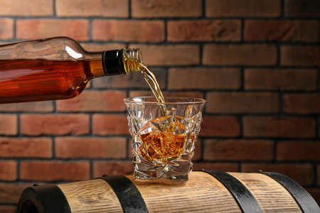 Pouring whiskey from bottle into glass on wooden barrel against brick wallの写真素材