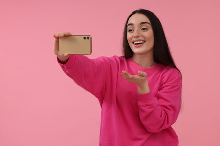 Smiling young woman taking selfie with smartphone on pink background, space for textの写真素材