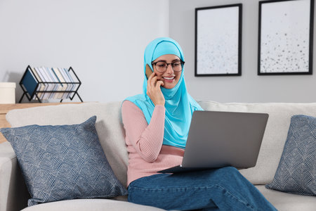 Muslim woman talking on smartphone near laptop at couch in room