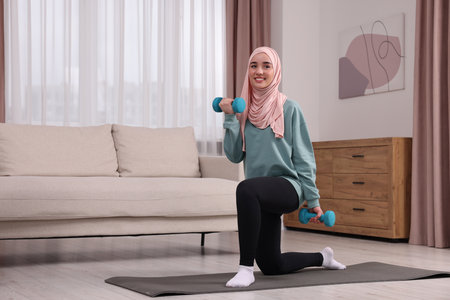 Muslim woman in hijab doing exercise with dumbbells on fitness mat at home. Space for textの写真素材