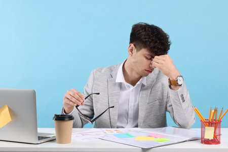 Tired young man working at white table on light blue background. Deadline conceptの写真素材