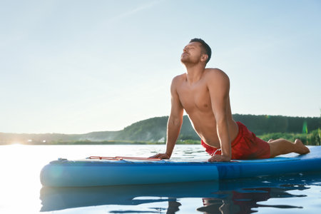 Man practicing yoga on light blue SUP board on river at sunsetの写真素材