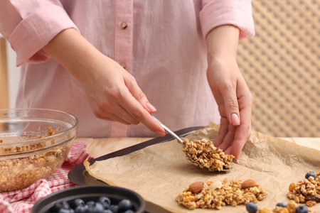Making granola bars. Woman putting mixture of oat flakes, dry fruits and other ingredients onto baking tray at table in kitchen, closeupの写真素材