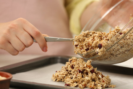 Making granola. Woman putting mixture of oat flakes, dried fruits and other ingredients onto baking tray, closeupの写真素材