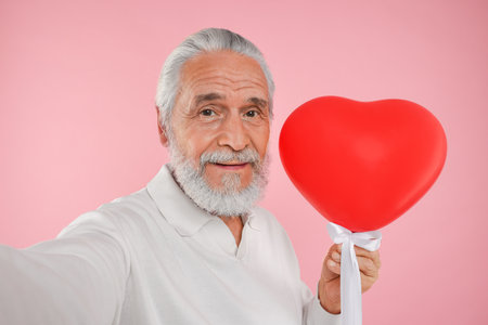 Senior man with red heart shaped balloon taking selfie on pink backgroundの写真素材