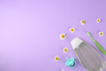 Bottle with baby oil, pacifier and toothbrush near daisies on violet background, flat lay. Space for textの写真素材