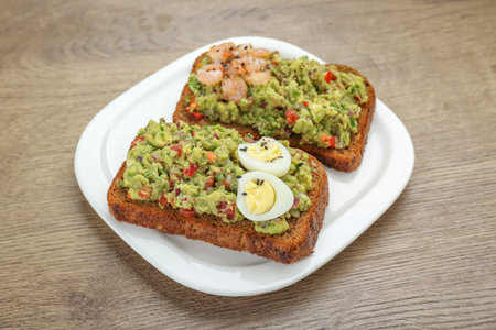 Slices of bread with tasty guacamole, eggs and shrimp on wooden table, closeup