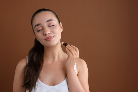 Beautiful young woman with roller bottle applying essential oil onto neck on brown background, space for textの写真素材