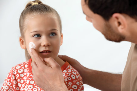 Father applying ointment onto his daughter's cheek on white backgroundの写真素材