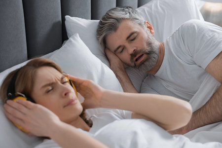 Irritated woman with headphones lying near her snoring husband in bed at homeの写真素材