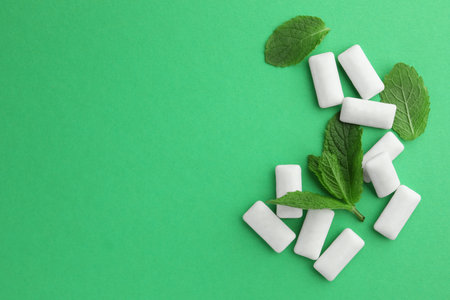 Tasty white chewing gums and mint leaves on green background, flat lay. Space for text