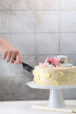 Woman cutting delicious cake decorated with macarons and marshmallows at white wooden table, closeup. Space for textの写真素材