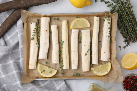Baking tray with raw salsify roots, lemon and thyme on white marble table, flat layの写真素材