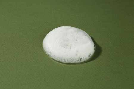 Sample of cleansing foam on dark green background, closeup. Cosmetic product