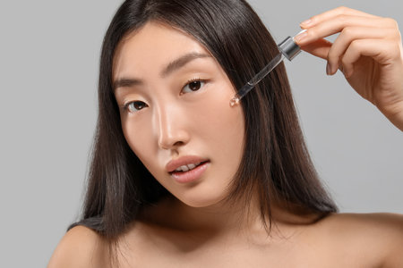 Beautiful young woman applying cosmetic serum onto her face on gray background, closeupの写真素材