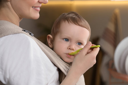 Mother feeding her cute child in sling (baby carrier) indoorsの写真素材
