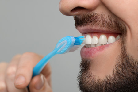 Man brushing his teeth with plastic toothbrush on light gray background, closeup