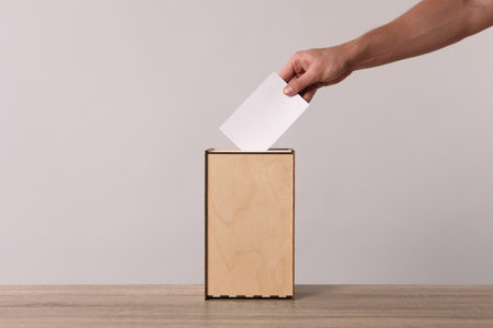 Man putting his vote into ballot box on light gray background, closeup