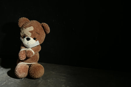 Stop child abuse. Tied toy bear with taped mouth and patches on grey floor against black background. Space for textの写真素材
