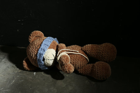 Stop child abuse. Tied toy bear with taped eyes lying on gray floor against black backgroundの写真素材