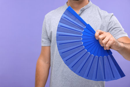 Man holding hand fan on purple background, closeup. Space for textの写真素材