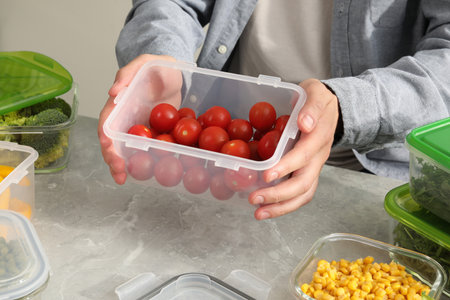 Man holding plastic container with fresh cherry tomatoes at light gray table in kitchen, closeup....