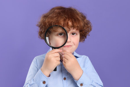 Cute little boy looking through magnifier glass on violet backgroundの写真素材