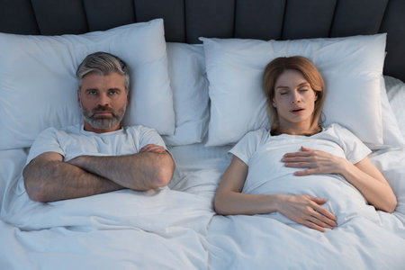 Irritated man lying near his snoring wife in bed at homeの写真素材