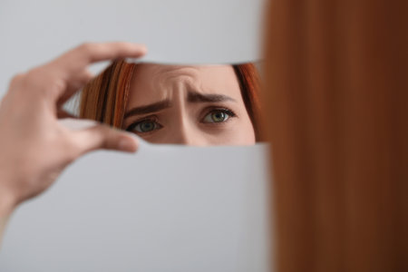 Young woman looking at herself in shard of broken mirror on light gray background, closeupの写真素材