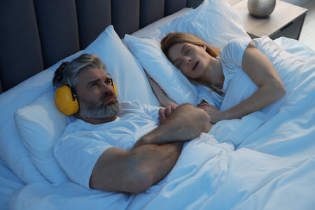 Irritated man with headphones lying near his snoring wife in bed at homeの写真素材