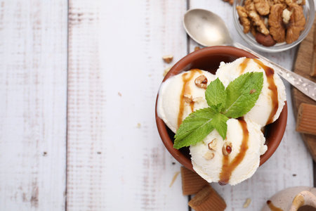 Bowl of tasty ice cream with caramel sauce, mint and nuts on white wooden table, flat lay. Space for textの写真素材
