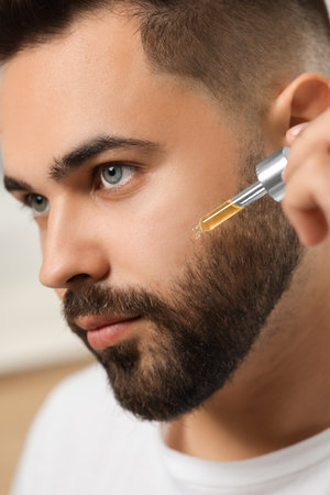Handsome man applying cosmetic serum onto his face on blurred background, closeupの写真素材