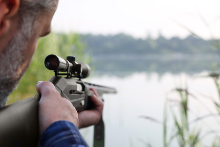 Man aiming with hunting rifle near lake outdoors, closeup. Space for textの写真素材
