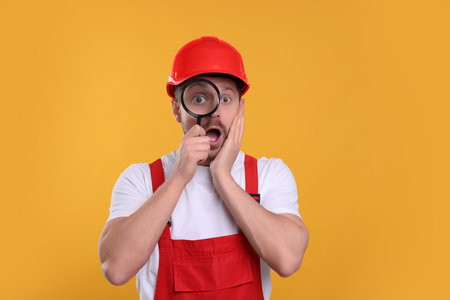 Surprised builder looking through magnifier glass on yellow backgroundの写真素材