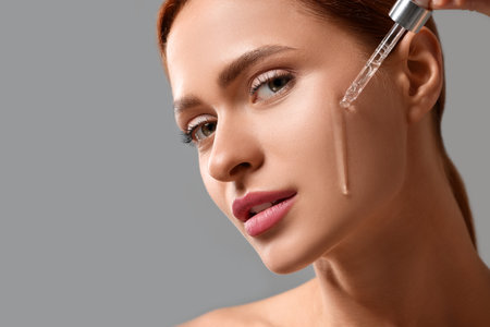 Beautiful young woman applying cosmetic serum onto her face on gray background, closeup. Space for textの写真素材